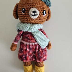 May include: A brown crocheted bear wearing a green and pink hat, a light blue scarf, and a red and pink plaid dress with yellow boots.