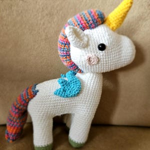 May include: A white crocheted unicorn with a yellow horn, blue wings, and a multi-colored tail. The unicorn is standing on a brown surface.