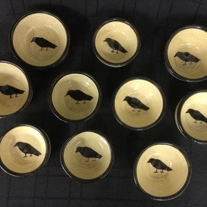 May include: Ten small ceramic bowls with black rims, each featuring a black silhouette of a crow on a cream-colored background.