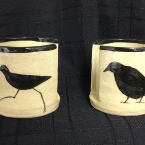 May include: Two ceramic mugs with black rim and black bird silhouettes. One mug features a bird with long legs, the other a crow.