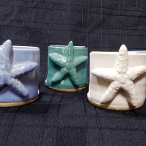 May include: Four ceramic starfish-shaped candle holders in different colors: blue, teal, white, and light blue. Each holder has a raised starfish design.