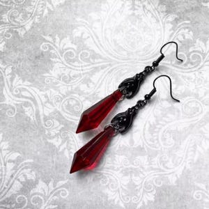 May include: A pair of gothic-style earrings featuring long, faceted, red crystal pendants. The earrings have black metal filigree accents and ear wires. The design is elegant and suitable for special occasions or everyday wear.