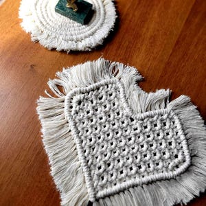 May include: Two off-white macrame coasters with fringe edges. One coaster is circular with a small green rectangular block topped with a gold-colored fish figurine. The other coaster is heart-shaped with a woven grid pattern and long fringe.