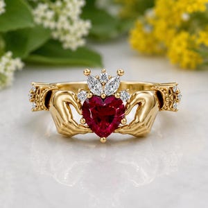 May include: A gold Claddagh ring with a heart-shaped, deep red gemstone at its centre, topped with a small diamond crown. The ring design includes two hands holding the heart, with diamond accents. The ring is a symbol of love.