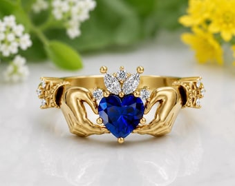 May include: A gold Claddagh ring with a heart-shaped, deep blue gemstone. The ring features two hands holding the heart, a crown above the heart, and small accent stones. The ring is set against a blurred floral background.