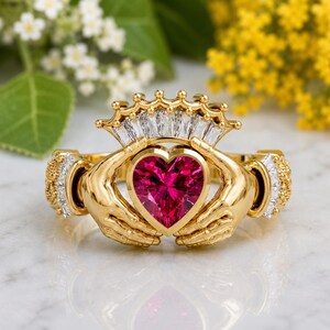 May include: A gold Claddagh ring with a heart-shaped, ruby-colored gemstone. The ring features two hands holding the heart, a crown above, and diamond accents on the sides. The ring is set against a blurred floral background.