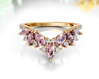 May include: A gold-colored ring with a V-shaped design. The ring features several pink marquise-cut gemstones and small, round, clear gemstones. The ring is set against a soft, blurred background with hints of white, blue, and yellow.