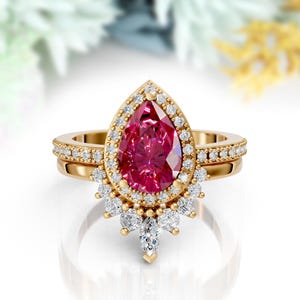 May include: A gold engagement ring set featuring a pear-shaped, vibrant pink gemstone surrounded by a halo of small, clear diamonds. The band is also adorned with small diamonds. A matching band sits below the main ring, creating a complete bridal set.