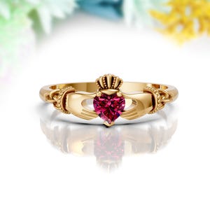 May include: A gold Celtic Claddagh ring with a red heart-shaped gemstone. The ring features two hands clasping the heart, symbolizing love, loyalty, and friendship.