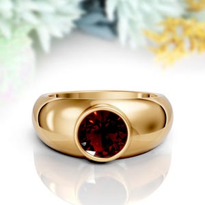 May include: A gold ring featuring a large, circular, deep red gemstone. The ring has a smooth, polished band and a modern design. The gemstone is the central focus, set within the gold band. The ring is a statement piece.