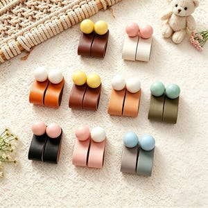 Può includere: Una collezione di anelli portatovaglioli decorativi in pelle. Ogni anello presenta una coppia di perline sferiche rotonde in vari colori, tra cui giallo, rosa, bianco e blu. Le fasce in pelle sono disponibili in tonalità marrone, arancione, nero e verde.