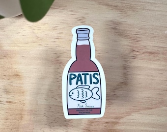 Może przedstawiać: Naklejka przedstawiająca butelkę sosu rybnego Patis. Butelka jest bordowa z białą etykietą z napisem "PATIS" nad ilustracją ryby i "Fish Sauce" poniżej. Tekst "PRODUCT OF THE PHILIPPINES" znajduje się na dole.