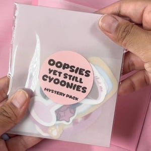 Puede incluir: Un paquete de plástico transparente que contiene pegatinas. Una pegatina redonda rosa en el paquete dice "OOPSIES YET STILL CYOONIES MYSTERY PACK" en texto negro. Las pegatinas del interior tienen varios colores y formas pastel.