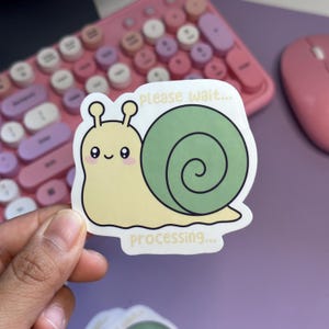 May include: A die-cut sticker featuring a light yellow snail with a green spiral shell. The snail has a smiling face with pink cheeks. The text "please wait..." is printed above the snail and "processing..." is printed below in yellow.