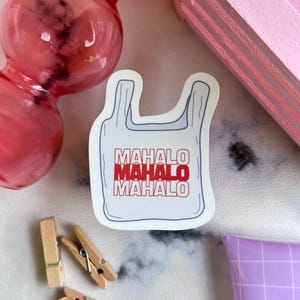 Puede incluir: Una pegatina blanca con forma de bolsa de la compra con la palabra "MAHALO" repetida tres veces en rojo y blanco. La pegatina está sobre una superficie blanca, con un objeto rosa y pinzas de madera en el fondo.