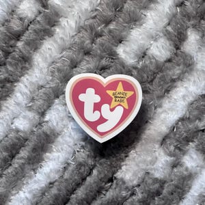 May include: A heart-shaped sticker with a pink background and a white border. The sticker features the word "ty" in white, a gold star with the words "Beanie Throwback Babe", and a white border.