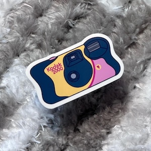 May include: A sticker featuring a retro Kodak 2000 camera design. The sticker has a yellow body, a dark blue lens area, and pink accents. The Kodak logo is in orange.