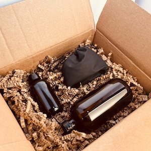 Peut inclure: Une boîte en carton remplie de papier de remplissage brun, contenant deux bouteilles en verre brun foncé et un sac noir à cordon. Les bouteilles sont de tailles et de formes différentes, suggérant un ensemble de produits.