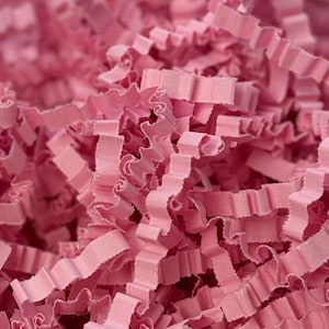 May include: Close-up of a pile of pink crinkle paper. The paper is cut into long, thin strips with a wavy, ruffled edge. The paper is a solid, light pink color and appears to be made of paper.