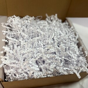 Puede incluir: Una caja de cartón marrón abierta, llena de papel triturado blanco. La caja está forrada con papel blanco. El papel triturado está bien embalado, proporcionando amortiguación.