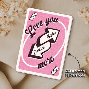 May include: A pink playing card with the words "Love you more" in a cursive font. Two white arrows point in opposite directions, each with a name. The text "NAMES CAN BE CUSTOM" is in a white box.