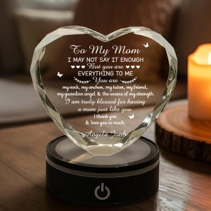 Puede incluir: Adorno de cristal en forma de coraz&oacute;n con el texto "To My Mom" y un mensaje de amor. El cristal transparente se asienta sobre una base negra con un bot&oacute;n de encendido iluminado. Una vela encendida est&aacute; en el fondo.
