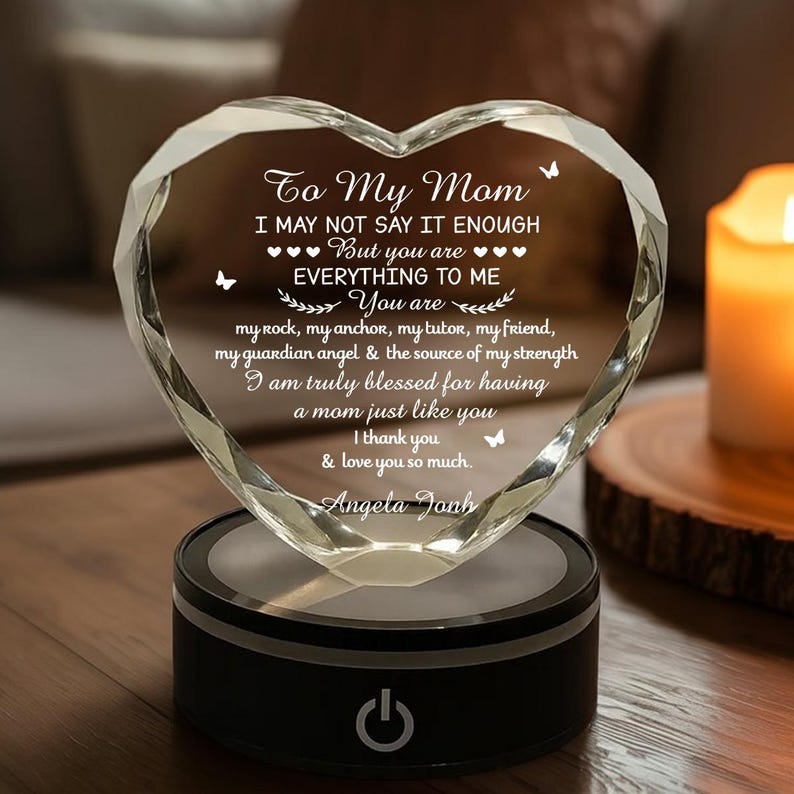 Puede incluir: Adorno de cristal en forma de coraz&oacute;n con el texto "To My Mom" y un mensaje de amor. El cristal transparente se asienta sobre una base negra con un bot&oacute;n de encendido iluminado. Una vela encendida est&aacute; en el fondo.