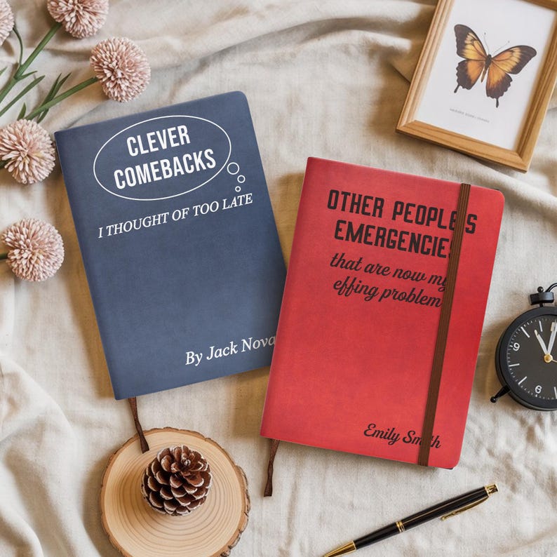Op de afbeelding: Twee hardcover notitieboeken met humoristische citaten. E&eacute;n is marineblauw met "CLEVER COMEBACKS" en "I THOUGHT OF TOO LATE". De andere is rood met "OTHER PEOPLES EMERGENCIES". Een ingelijste vlinder en een zwarte klok zijn ook zichtbaar.