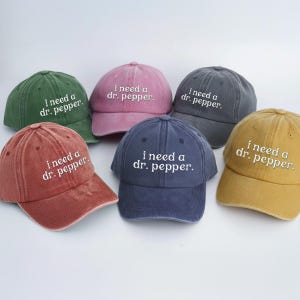 May include: Six baseball caps in various colors, including green, pink, and navy blue. Each cap has the phrase "i need a dr. pepper." embroidered in white. The caps are arranged on a white surface.