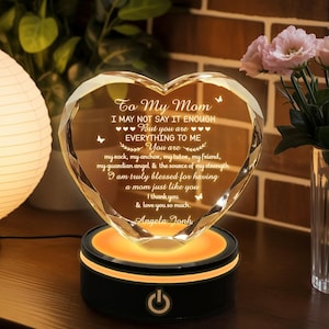 Puede incluir: Un adorno de cristal en forma de coraz&oacute;n con el texto "To My Mom" y un mensaje de amor, iluminado por un c&aacute;lido resplandor naranja desde su base negra. El adorno es un regalo reflexivo.