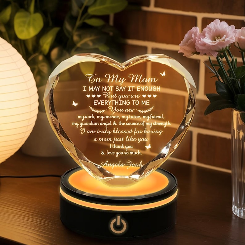Puede incluir: Un adorno de cristal en forma de coraz&oacute;n con el texto "To My Mom" y un mensaje de amor, iluminado por un c&aacute;lido resplandor naranja desde su base negra. El adorno es un regalo reflexivo.