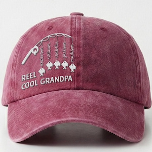 May include: A maroon baseball cap with a curved brim. The cap features white embroidered text and a fishing-themed design. The text reads "REEL COOL GRANDPA" and includes names hanging from fishing lines.