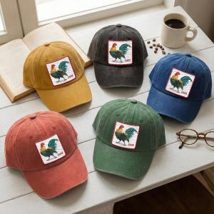 May include: Six baseball caps in various colors: mustard yellow, charcoal, navy blue, green, and rust. Each cap features a square patch with a rooster illustration and the word "COCK".