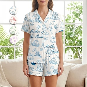 May include: White pajama set with a blue wedding-themed pattern, including cakes, bells, and cars. The words "Mother of the Bride" are printed on the shirt. The set includes a short-sleeved top and shorts.