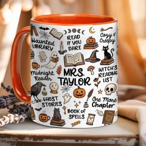 May include: A white ceramic mug with an orange interior and handle, featuring a repeating pattern of Halloween-themed illustrations including ghosts, black cats, pumpkins, skulls, and books, with the text Mrs. Taylor centred on the front.