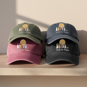 May include: A stack of six baseball caps in various colors: olive green, dusty rose, charcoal gray, and black. Each cap features an embroidered design of figures, a sun, and a dog, with the text "Walk for Peace".