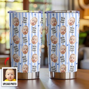 May include: Two stainless steel tumblers with a custom design. The tumblers feature a repeating pattern of a baby's face, hearts, and the text "Best Dad Ever" on a blue and white striped background. The bottom of the tumblers are silver.