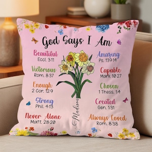 May include: A pink decorative pillow with the text "God Says I Am" and inspirational words. A floral bouquet with yellow and white flowers is in the center. The pillow has a floral border.