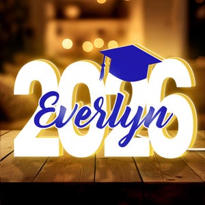 May include: A lighted graduation decoration featuring the year "2026" in white, with the name "Everlyn" in blue script. A blue graduation cap sits atop the numbers. The sign is on a wooden surface.