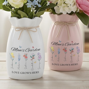 May include: Two ceramic vases, one white and one pink, each adorned with floral designs and the text "Mom's Garden" and "Love Grows Here." The vases are tied with twine and hold colorful flowers, creating a charming, decorative display.