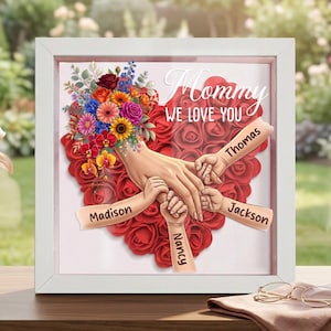 May include: A framed artwork with a white border features a heart made of red roses. A hand and three smaller hands are joined together, with the names "Madison", "Thomas", "Jackson", and "Nancy" on the arms. The words "Mommy WE LOVE YOU" are written above.