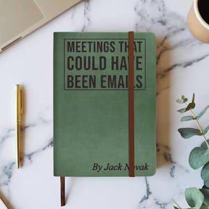 Op de afbeelding: Een groen notitieboek met de tekst "MEETINGS THAT COULD HAVE BEEN EMAILS" in het zwart. Het notitieboek heeft een bruine elastische band en de naam van de auteur, "By Jack Novak", onderaan. Een gouden pen en een laptop zijn zichtbaar op de achtergrond.