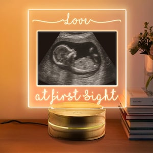 May include: A light-up frame with the words "love" and "at first sight" in cursive. The frame shows a grayscale ultrasound image of a foetus. The base is made of wood.