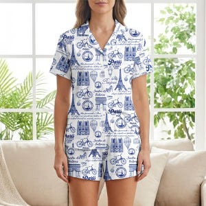 以下が含まれることがあります： パリをテーマにした青いプリントの白いパジャマセット。半袖トップスとショーツには、エッフェル塔、自転車、その他のパリのランドマークのイラストが描かれています。「Paris is always a good idea」というテキストも表示されています。