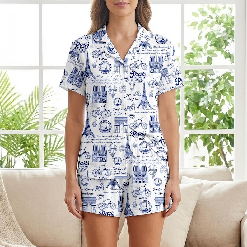 以下が含まれることがあります： パリをテーマにした青いプリントの白いパジャマセット。半袖トップスとショーツには、エッフェル塔、自転車、その他のパリのランドマークのイラストが描かれています。「Paris is always a good idea」というテキストも表示されています。