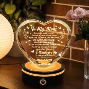 May include: A heart-shaped crystal ornament with the text "To My Mom" and a loving message, illuminated by a warm, orange glow from its black base. The ornament is a thoughtful gift.