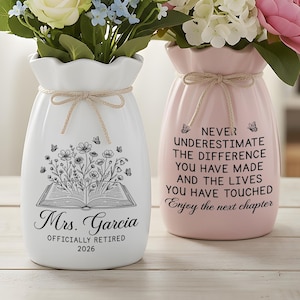 May include: Two ceramic vases, one white and one light pink, each tied with a tan twine bow at the neck. The white vase features a black line drawing of an open book with flowers and butterflies, plus the text Mrs. Garcia OFFICIALLY RETIRED 2026. The pink vase displays the text NEVER UNDERESTIMATE THE DIFFERENCE YOU HAVE MADE AND THE LIVES YOU HAVE TOUCHED Enjoy the next chapter.