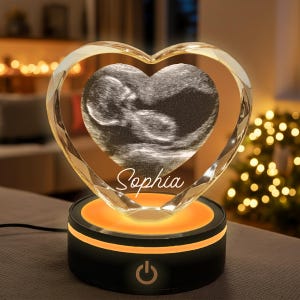 May include: A heart-shaped crystal keepsake with a sonogram image and the name "Sophia." The crystal sits on a black base with an orange glow, and a power button. The heart is faceted, reflecting light.