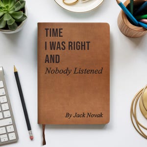 Op de afbeelding: Een bruin notitieboek met de tekst "TIME I WAS RIGHT AND Nobody Listened" en "By Jack Novak" gedrukt op de kaft. Het notitieboek heeft een lint als bladwijzer. Het notitieboek ligt op een wit oppervlak met een potlood, toetsenbord en andere kantoorbenodigdheden.