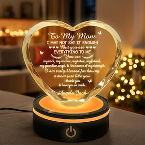 Puede incluir: Adorno de cristal en forma de coraz&oacute;n con el texto "To My Mom" y un mensaje de amor. El cristal transparente se asienta sobre una base negra con un brillo naranja y un bot&oacute;n de encendido. Un regalo reflexivo.